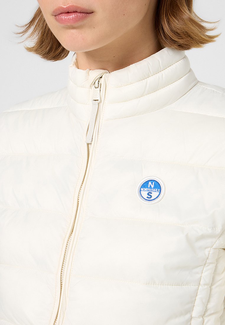 Giacca imbottita bianca con colletto alto, dotata di un logo circolare blu sul petto e una cerniera frontale. Texture liscia con dettagli cuciti.