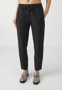 Schwarze, schmal geschnittene Hose mit elastischem Zugbandbund, Seitentaschen und glatter Textur. Kombiniert mit gemusterten Schlangenleder-Schuhen.