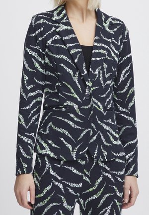 Femme portant un blazer noir et un pantalon avec un motif à rayures florales blanches et vert clair, et un top noir en dessous.