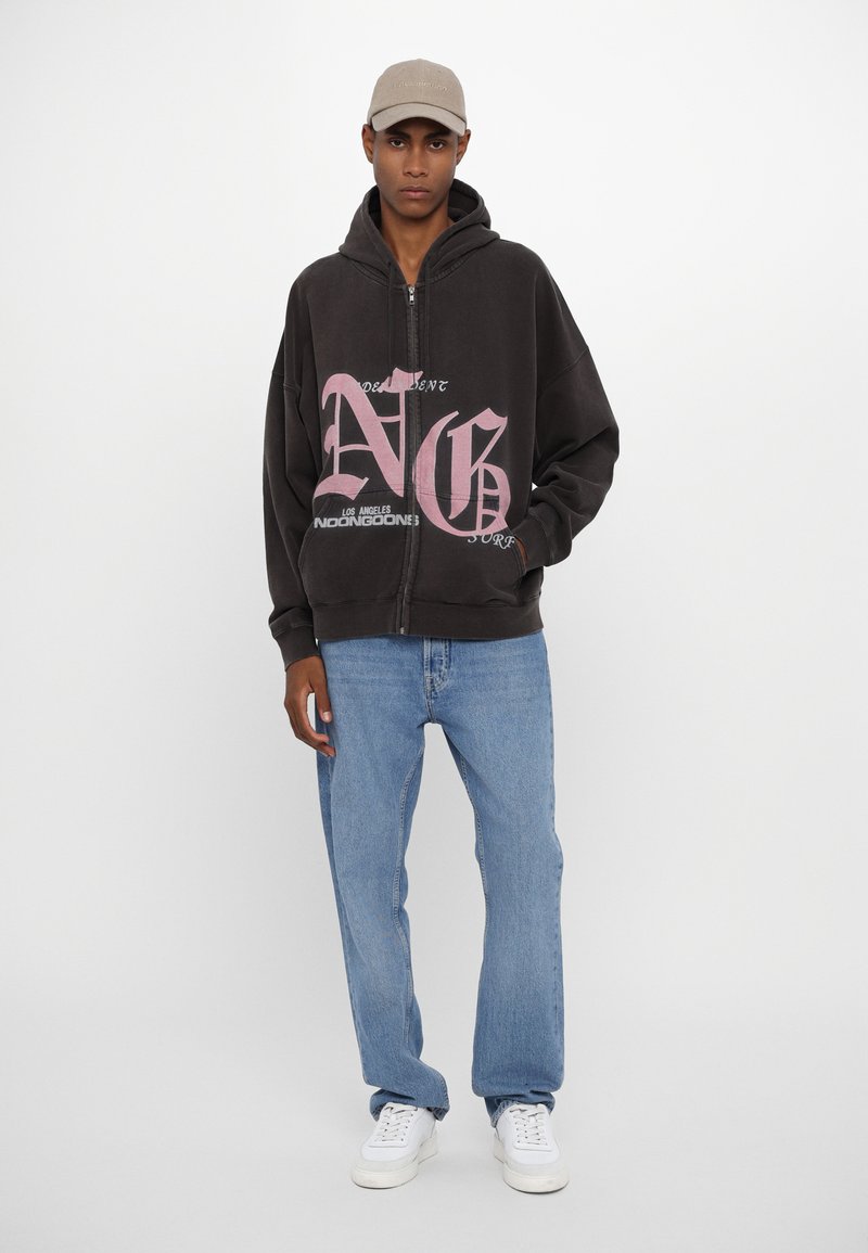 Haut à capuche zippé noir avec un texte graphique rose, associé à un jean bleu clair et des baskets blanches. Le chapeau est beige, ajoutant une touche neutre.