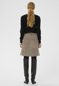 Brauner Wildlederrock in A-Linie mit zwei Gesäßtaschen, kombiniert mit enganliegenden schwarzen Overknee-Stiefeln und einem schwarzen Pullover, von hinten gesehen.