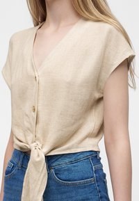Femme portant un haut beige à manches courtes avec col en V, boutons et nœud devant, associé à un jean en denim bleu.