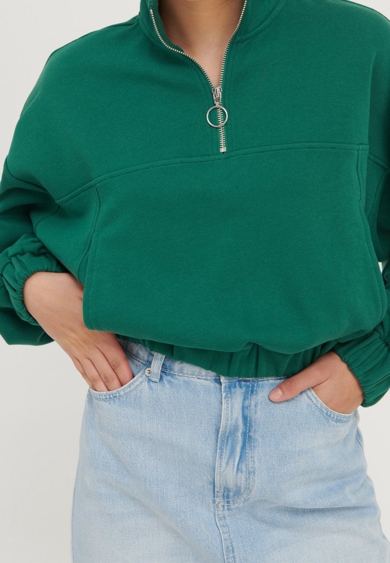 Sudadera de forro polar verde con cremallera y cuello alto, puños elásticos y un bolsillo frontal. Combinada con pantalones cortos de mezclilla de color azul claro.