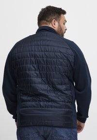 Tamsiai mėlynas švarkas su šiuolaikišku siuvimu nugarinėje dalyje, minkštomis medžiagos rankovėmis ir aukštu apykakle. Jis turi tekstūruotą paviršių ir laisvą siluetą.