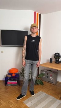 Čierna bezrukávová košeľa s grafikou "CELESTIAL", svetlosivé rozšírené džínsy, čierne slip-on topánky a piesková plochá čiapka. Moderné vnútorné prostredie.