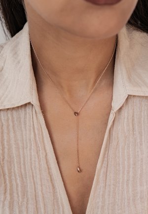 Rose gold necklace with a heart-shaped pendant, featuring a fine chain and a teardrop accent, set against a textured beige shirt.