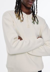 Cremfarbener Strickpullover mit Henley-Kragen, der drei Knöpfe, gerippte Bündchen und eine weiche Textur aufweist.