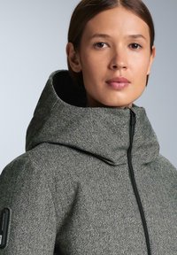 Sivá bunda s kostkovaným vzorom houndstooth, ktorá má vysoký golier, predný zips a bočné vrecko s čiernymi kovovými detailmi.