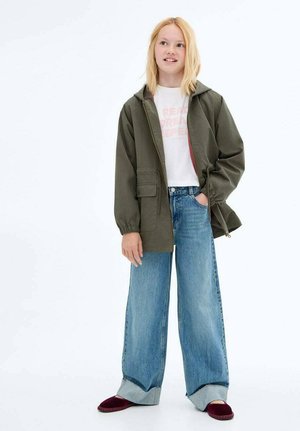 Giacca cappuccio verde oliva con zip, abbinata a una t-shirt grafica bianca e jeans in denim blu larghi, con gambe ampie e orli arrotolati.