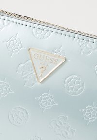 Pochette gaufrée bleu clair avec un logo triangulaire doré. La surface texturée présente des motifs floraux et géométriques. Fermeture à glissière sur le dessus.