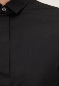 Chemise noire à col avec patte boutonnée et logo A|X discret brodé sur la poitrine.