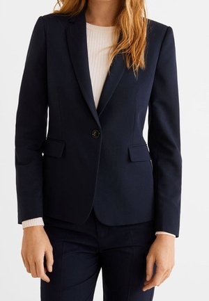 Kvinde iført en skræddersyet mørk navy blazer med knaplukning foran, kombineret med matchende bukser og en lys ribbet sweater nedenunder.