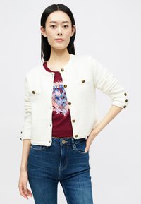 Cardigan branco texturizado com bolsos frontais, botões dourados e mangas três quartos, sobre uma t-shirt gráfica em bordô e jeans azuis.