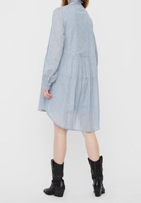 Abito blu chiaro a maniche lunghe realizzato in un tessuto morbido e trasparente. Caratterizzato da una vita arricciata e un orlo curvo. Stivali neri con tacco completano l'outfit.