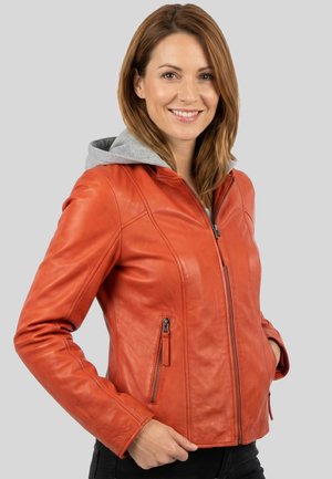 Lächelnde Frau mit glatten hellbraunen Haaren, die eine orange Lederjacke mit grauer Kapuze und schwarze Hose trägt, Hände in den Taschen.