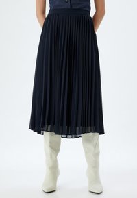 Falda midi plisada de color marino con cinturilla ajustada y bolsillos, combinada con botas de cuero blanco hasta la rodilla. Tela suave y texturada.