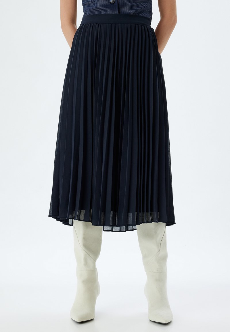 Falda midi plisada de color marino con cinturilla ajustada y bolsillos, combinada con botas de cuero blanco hasta la rodilla. Tela suave y texturada.