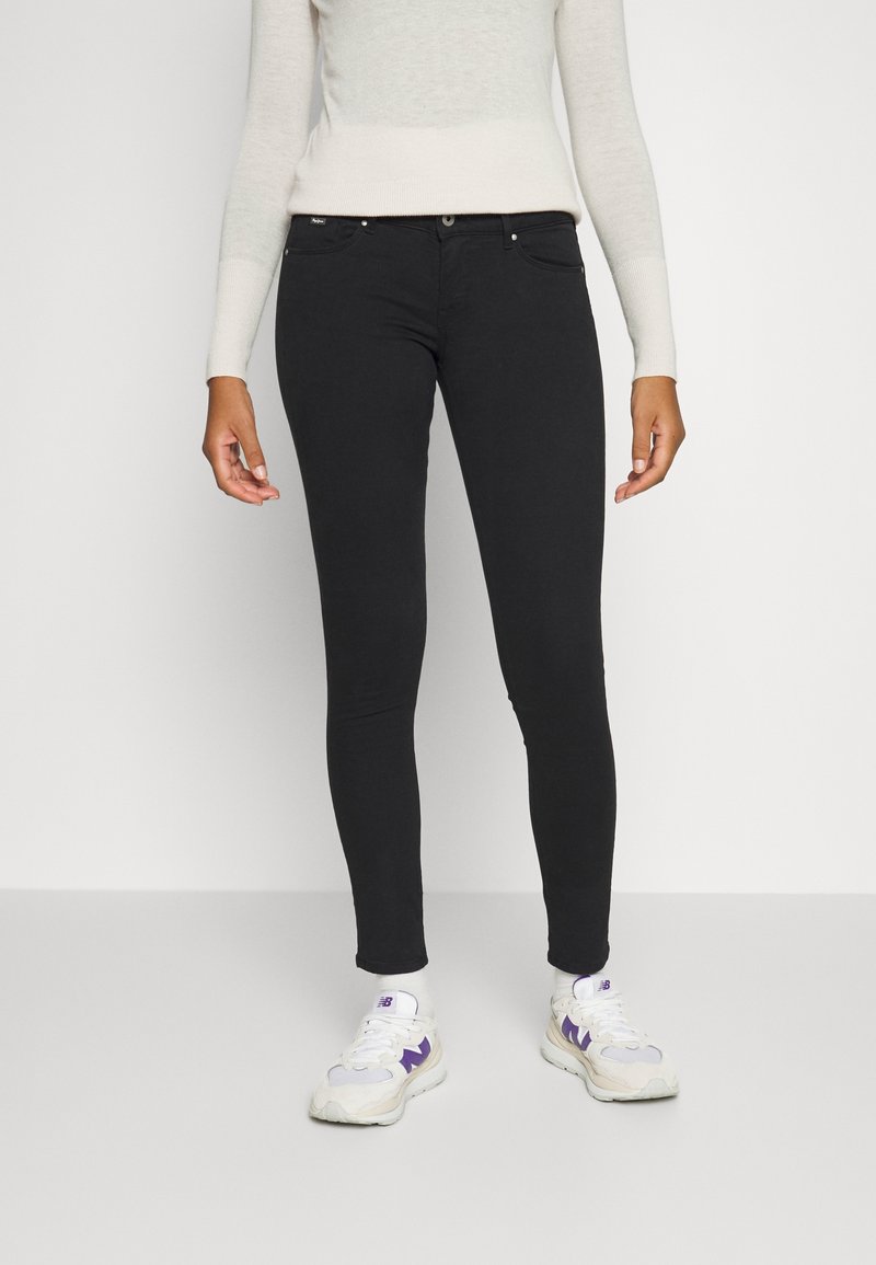 Jeans slim fit negros con una textura suave, que cuentan con bolsillos frontales y cierre de botón metálico. Combinados con zapatillas blancas con acentos morados.