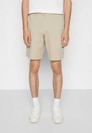 Homme portant un short beige jusqu'aux genoux, des chaussettes blanches et des baskets blanches, debout devant un fond uni.
