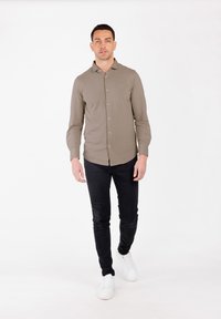Camicia a maniche lunghe marrone chiaro con colletto a bottoni, abbinata a pantaloni neri slim-fit e sneakers bianche, in piedi davanti a uno sfondo bianco.