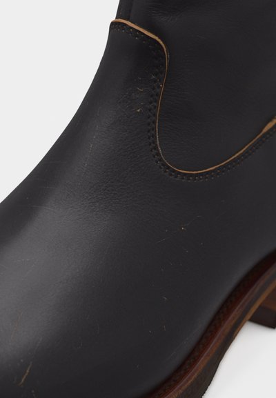 Κοντινό πλάνο μαύρης δερμάτινης μπότας αστραγάλου με καφέ ραφές και ξύλινη σόλα, που δείχνει την υφή και σημάδια φθοράς στην επιφάνεια.