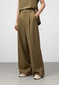 Person som bär vida olivgröna byxor och matchande topp, med en hand i fickan och vita sneakers med beige detaljer.