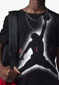 Czarny t-shirt z białym konturem sylwetki jump man w efekcie rozprysku, z czerwonym tekstem "AIR" poniżej, noszony z czarnym plecakiem i nałożonymi łańcuchami.