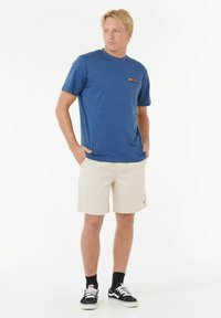 Blaues Rundhals-T-Shirt mit rotem Logo, kombiniert mit beigen Shorts. Das Modell trägt schwarz-weiße Sneakers mit schwarzen Socken. Einfaches Design, lässiger Stil.