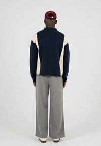 Granatowy sweter z kremowymi panelami bocznymi, z kołnierzem. W połączeniu z jasnoszarymi spodniami o szerokich nogawkach. Model odwrócony plecami, noszący bordową czapkę.