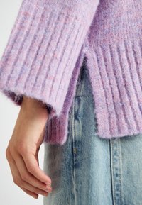 Svijetlo ljubičasti rebrasti pulover s mekanom, pahuljastom teksturom, s bočnim prorezom i širokim rukavima, nosi se preko svijetloplavih traperica.