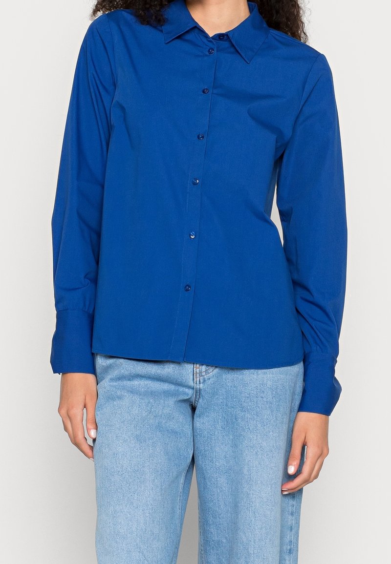 Blaues Langarmhemd mit Kragen, Knopfleiste und Manschettenakzenten, aus glattem Stoff. Kombiniert mit hellblauen Jeans.