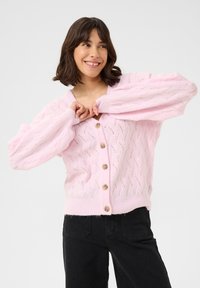 Cardigan en tricot rose avec un col en V, un motif de câbles texturé et de grands boutons. Présente des manches bouffantes et un ourlet côtelé. Associé à un pantalon noir.