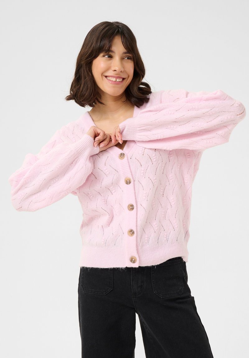 Cardigan en tricot rose avec un col en V, un motif de câbles texturé et de grands boutons. Présente des manches bouffantes et un ourlet côtelé. Associé à un pantalon noir.