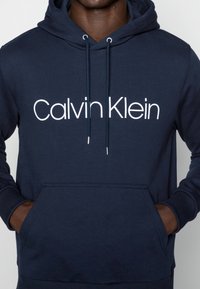 Man som bär en marinblå hoodie med "Calvin Klein"-logotyp i vitt tryckt på bröstet, händerna i fickan fram, inget synligt ansikte.
