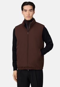 Hnedý puff vest s vysokým golierom, predným zipsom a bočnými vreckami, vrstvený cez čierny rolák, s ľahkou textúrou a čistými líniami.