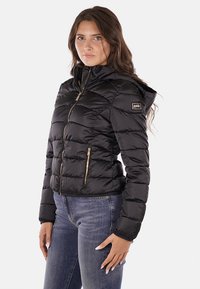Giacca a vento nera con cappuccio, dotata di chiusura con zip, texture trapuntata e tasche con zip con dettagli in tonalità dorata.