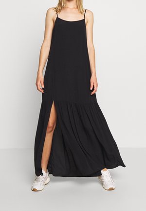 Kobieta ubrana w czarną, bezrękawnikową sukienkę maxi z bocznym rozcięciem, połączoną z białymi trampkami, stojąca na jednolitym jasnym tle.