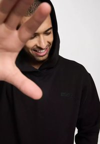 Czarna bluza z kapturem o teksturowanej tkaninie, z małą czarną łatą logo z przodu. Model jest częściowo zasłonięty przez uniesioną dłoń.