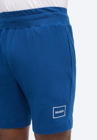 Μπλε βερμούδα μέχρι το μέσο του μηρού με ελαστική μέση και πλαϊνές τσέπες, με λευκό τετράγωνο λογότυπο που αναγράφει "HUGO" στο αριστερό πόδι.