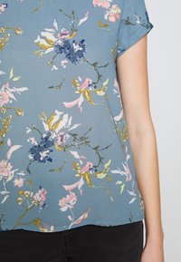 Blusa floral en azul con acentos en rosa, blanco y dorado. Mangas cortas, corte holgado, textura suave y diseño contemporáneo.
