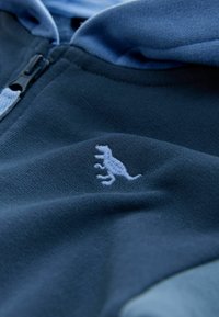 Nahaufnahme eines marineblauen Reißverschlusskleidungsstücks mit hellblauer Dinosaurier-Stickerei in der Nähe des Reißverschlusses.