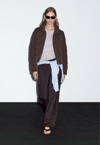 Donna che cammina con occhiali da sole, giacca trapuntata marrone, maglia grigia, pantaloni larghi neri, maglione azzurro chiaro legato in vita e sandali neri.