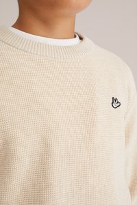 Beige tekstūrēts džemperis ar ribotu apkakli un mazu melnu izšūtu logo uz krūtīm, virs balta krekla apkakles.