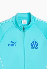 Chaqueta azul claro con cremallera, hecha de tela lisa. Presenta el logo de Puma, el texto "KING" y el emblema de Marsella en azul oscuro. Incluye detalles con patrones.