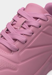 Gros plan sur une basket rose avec des lacets épais, un matériau synthétique texturé, et des détails de panneaux géométriques sur le côté.