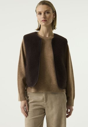Woman wearing brown knit sweater, dark brown sleeveless textured vest, and beige corduroy pants standing against white background.