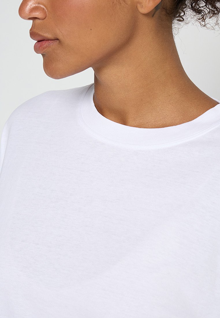 Nahaufnahme eines Halses und des unteren Gesichts einer Person, die ein schlichtes weißes Rundhals-T-Shirt trägt, vor einem neutralen Hintergrund.