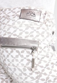 Jeans blancos con un sutil patrón de hojas en gris claro, con un bolsillo metálico con cremallera y una etiqueta de marca texturizada en la cinturilla.