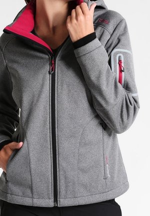 Femme portant une veste softshell grise zippée avec une doublure rouge, une poche zippée sur le bras et des détails noirs aux poignets, les mains près du col et de la poche.