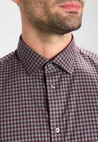 Camicia a quadri in tessuto marrone e grigio con colletto a punta, caratterizzata da due bottoni grigi e una leggera trama.
